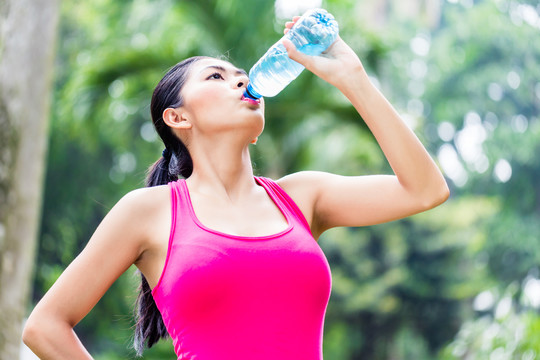 一名亚洲女子在热带公园的运动训练中休息，饮用瓶装水