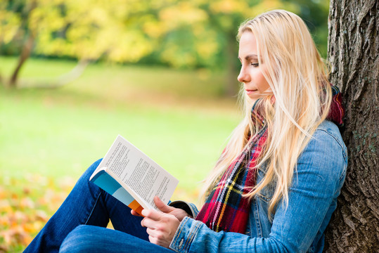 在秋天的公园里，坐在绿叶覆盖的草地上看书的女人