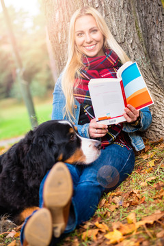 一位妇女和她的伯尔尼山狗在秋天公园里看书，坐在长满树叶的草地上