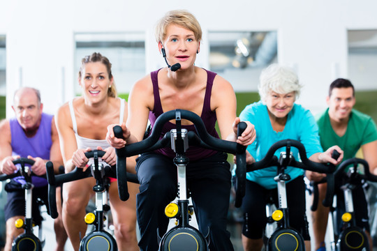 老年人和年轻人在健身房骑健身自行车进行耐力和有氧运动训练，教练正在引导他们