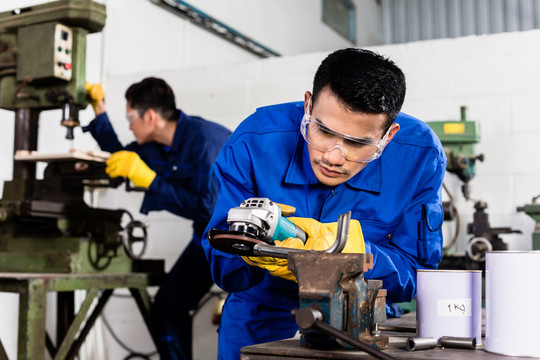 两名亚洲工业工人在金属厂使用电动磨具和电动钻床