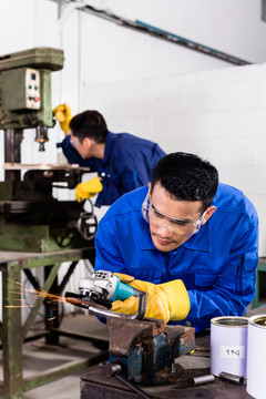 两名亚洲工业工人在金属厂使用电动磨具和电动钻床