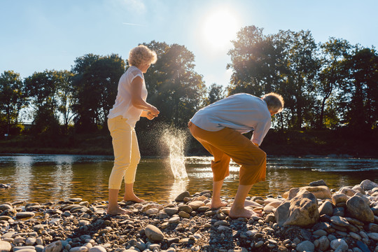 在阳光明媚的日子里，两位赤脚老人一边享受退休生活，一边向河里扔石头，这是一幅完整的后视图