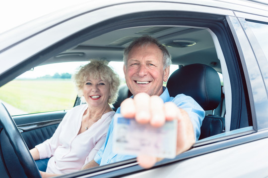 一位快乐的老人坐在他快乐的妻子旁边，展示他现有的驾驶执照