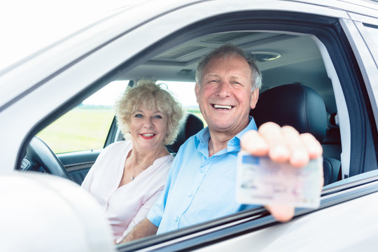 一位快乐的老人坐在他快乐的妻子旁边，展示他现有的驾驶执照