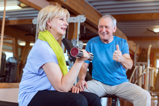 一位年长的女士在健身房练习哑铃健身，而她的丈夫则让她兴奋起来