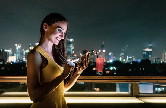 美丽的女人在俯瞰城市天际线的屋顶酒吧检查她的手机
