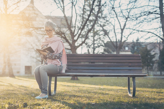 一位年长的女士独自坐在公园的长椅上，在傍晚的阳光下看书