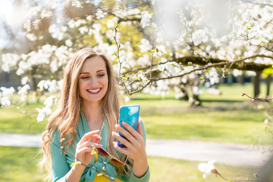 在春天的一个美丽的日子里，一个年轻的女人站在一棵盛开的树上用她的手机