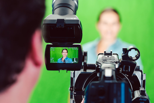 摄像机指向绿屋演播室里的一名女记者或记者，镜头对准了控制监视器