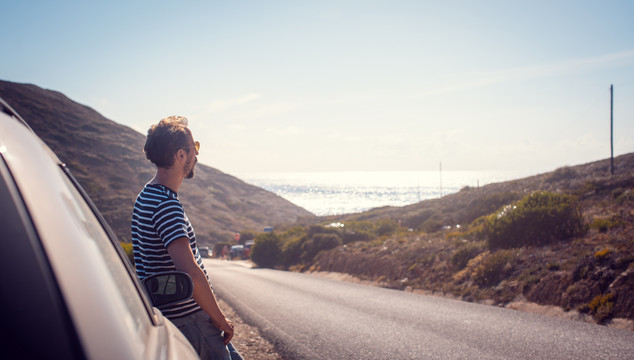 在公路旅行中，一名男子坐在汽车旁休息，看着远处的大海