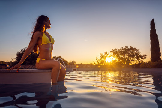 美丽的女人坐在靠近水面的游泳池上看日落
