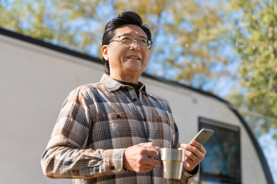 一个老年男人站在房车前拿着手机喝咖啡