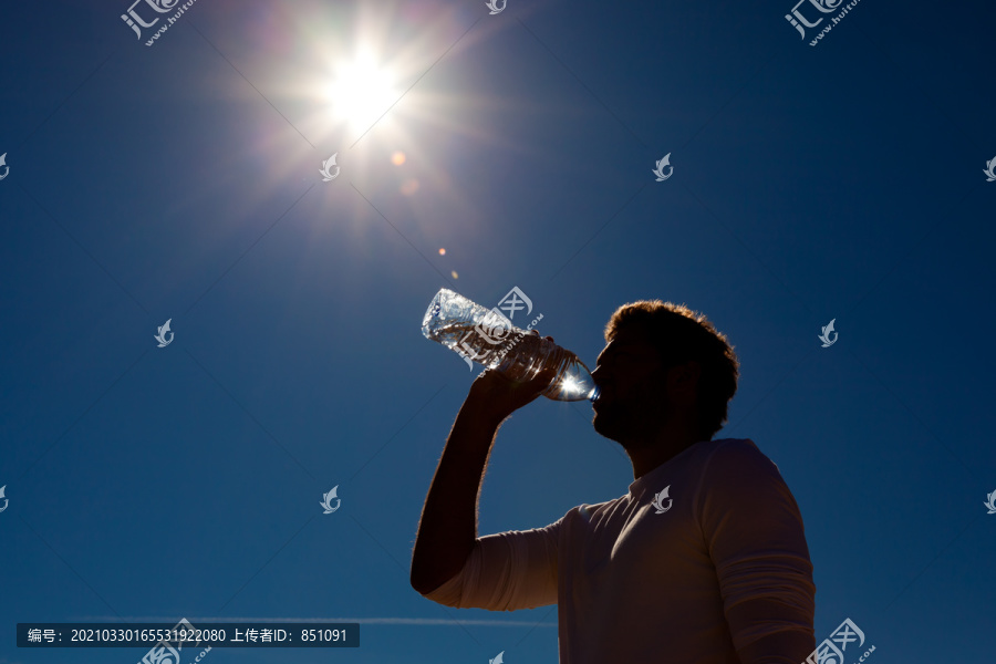 在炎热的阳光下，在蓝天的背景下，一个喜欢运动的男人从瓶子里喝水