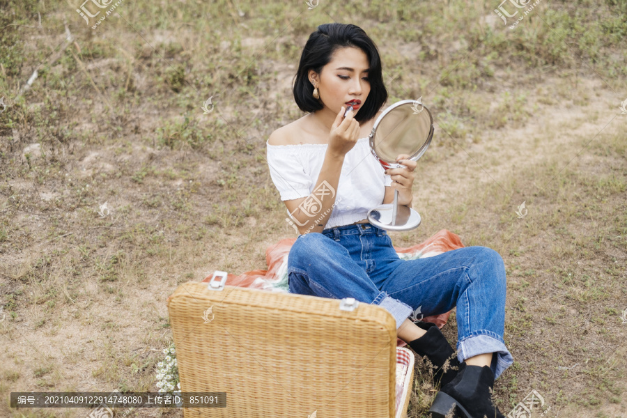 年轻性感的亚洲女孩正在野餐，坐在垫子上，化妆，拿着一个便携式镜子，享受化妆的乐趣。