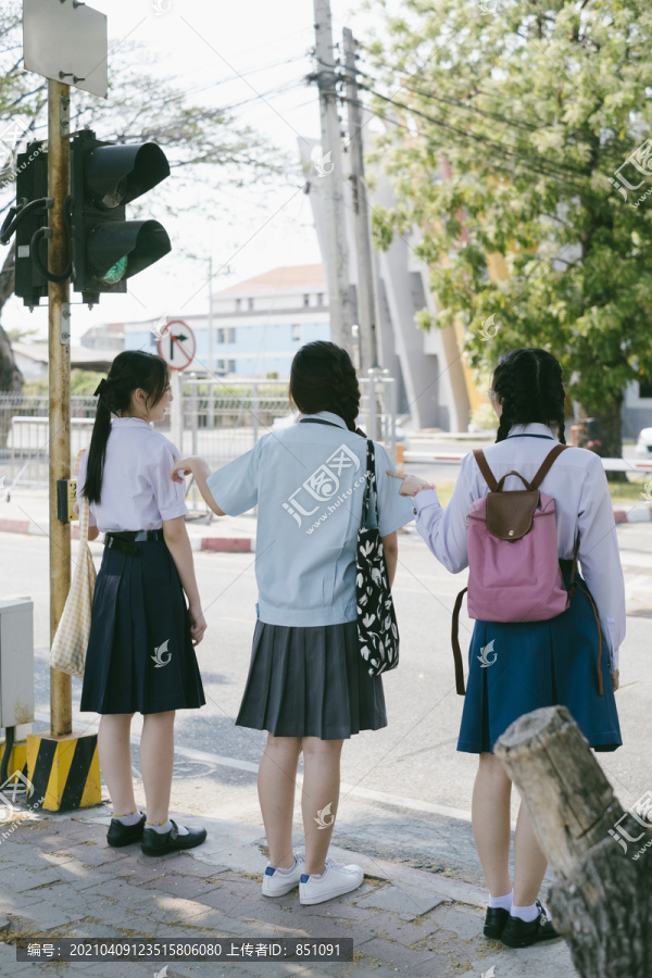 三个穿着不同制服的漂亮亚泰学生在过马路前推着红绿灯杆。