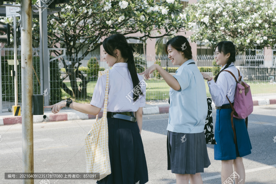 三个穿着不同制服的漂亮亚泰学生在过马路前推着红绿灯杆。