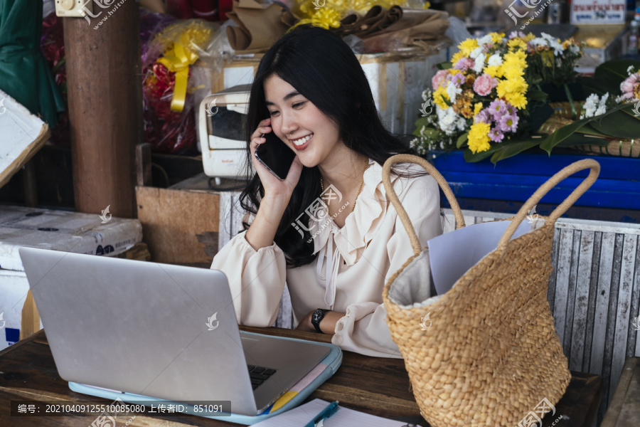 穿着休闲衬衫的泰国工作女商人在电话里谈笑风生。在家里用笔记本电脑工作。