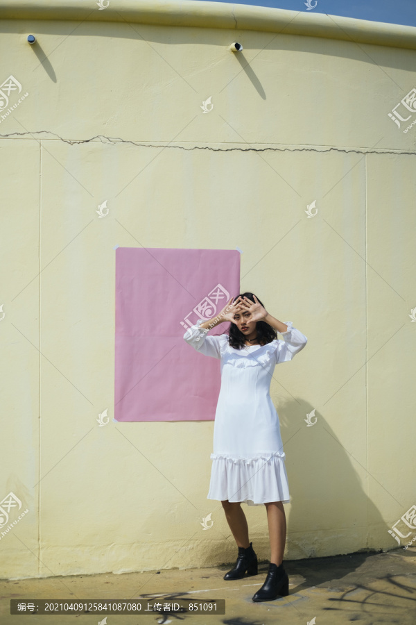黑色卷曲的长发女孩身着白色连衣裙，以别致的姿态站在黄色的墙边，用粉色的纸，复制空间。