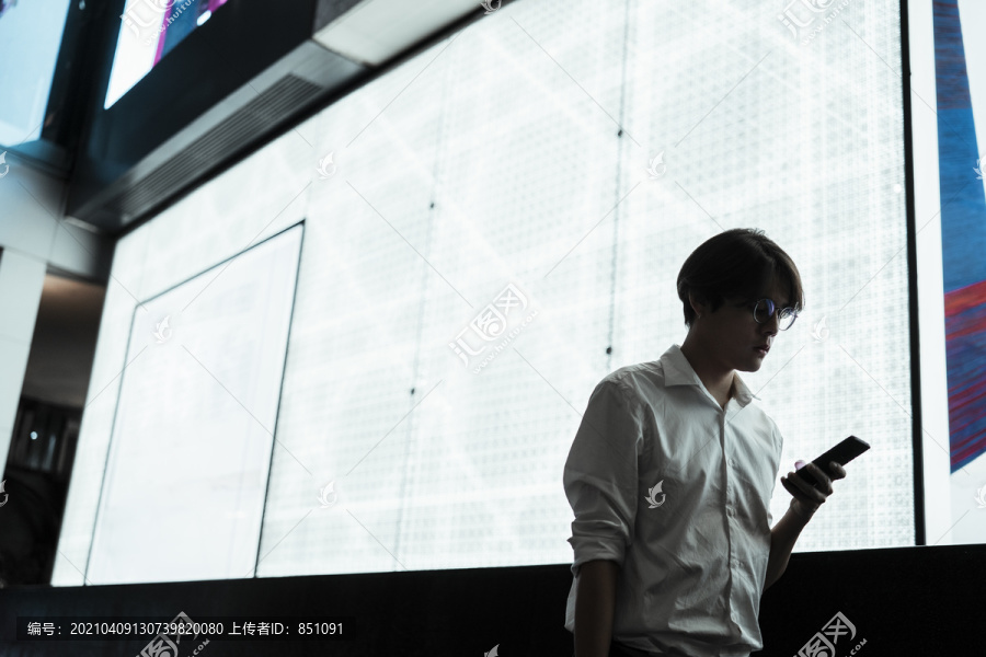 白光下，穿着白衬衫的人拿着手机站在商场广告牌前的剪影。
