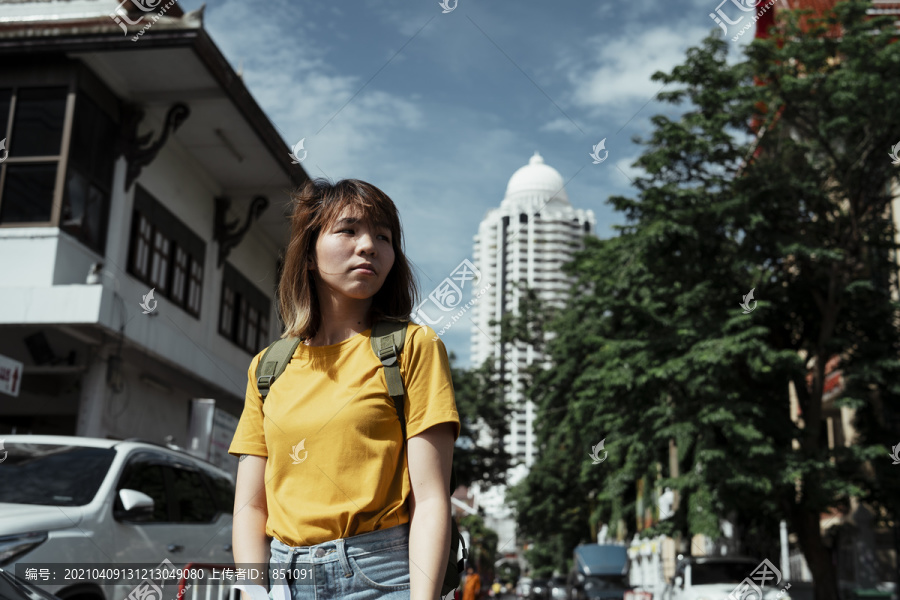 黄色的背包女，背着绿色的背包，站在以庙宇、树木和高楼为背景的城市里。