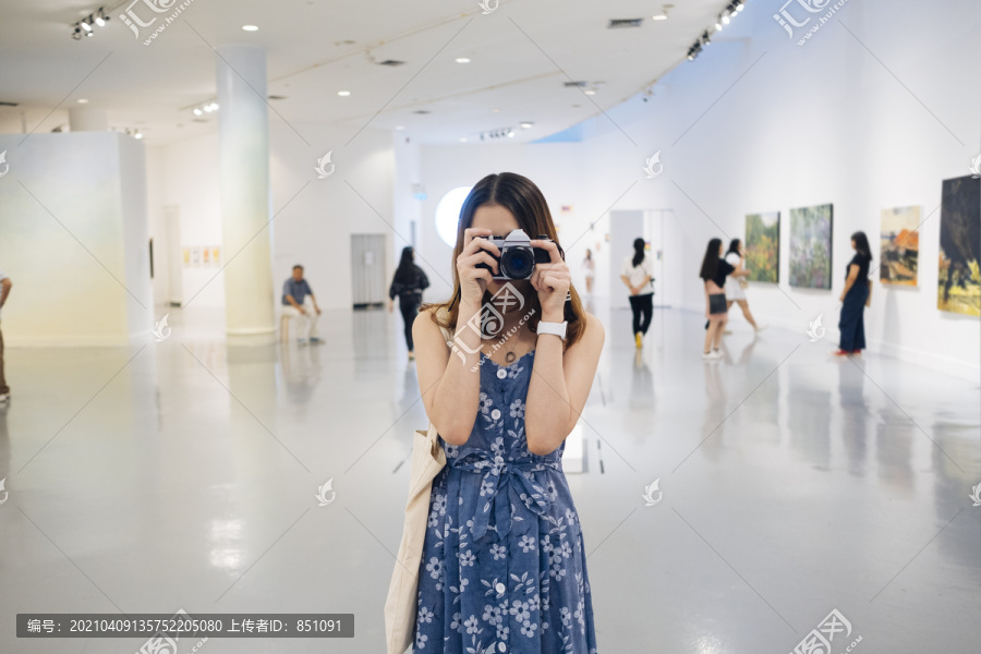 在美术馆里，一位身着蓝色连衣裙的泰国亚裔年轻女子正在用相机拍照。