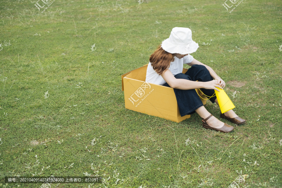 年轻的泰国亚裔女子留着时髦的卷发，穿着白衬衫，戴着帽子，坐在纸箱里，手里拿着黄色的玩具扩音器。