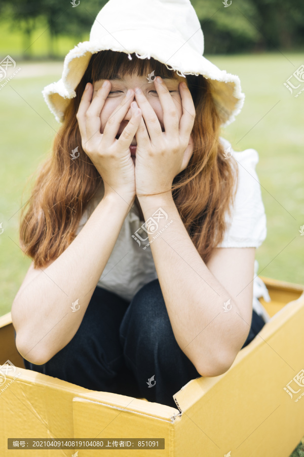 在大自然的绿色草地上，嬉皮的泰国亚裔年轻女子留着时髦的卷发，穿着白衬衫，戴着帽子，坐在纸板箱里。