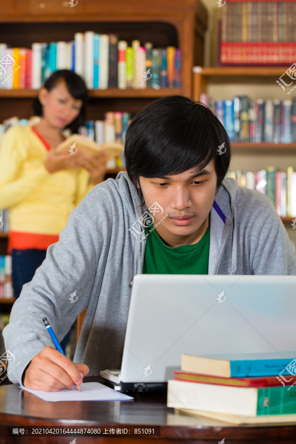 学生-年轻的亚洲男子在图书馆用笔记本电脑学习，一名女学生站在背景书架上看书