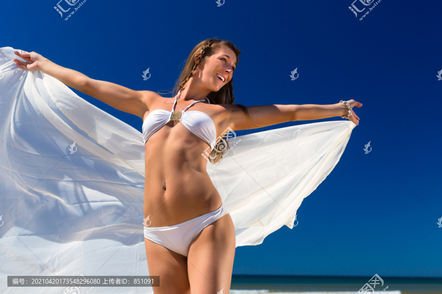 一个穿着白色比基尼的漂亮女人站在沙滩上晒太阳，戴着一条白色的围巾，天空一片蔚蓝