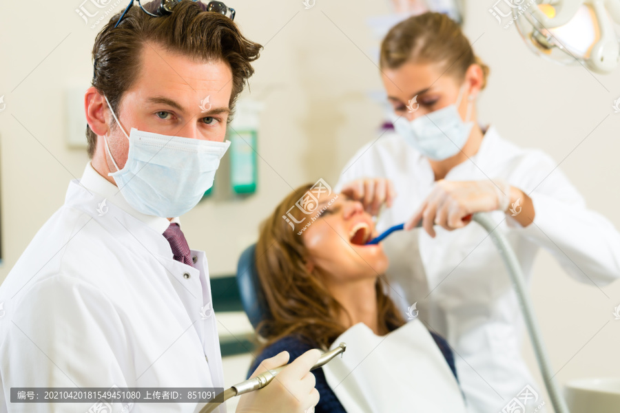 牙医在他的手术中看着观看者，背景是他的助手正在给一名女性患者治疗