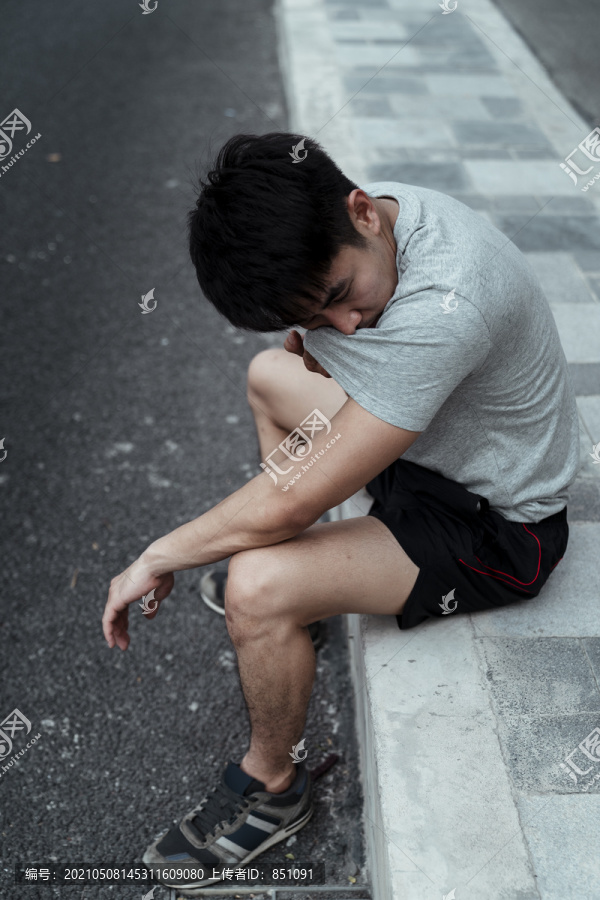 运动后在人行道上休息的疲倦的人。亚洲年轻人用衬衫擦脸。跑步后休息。