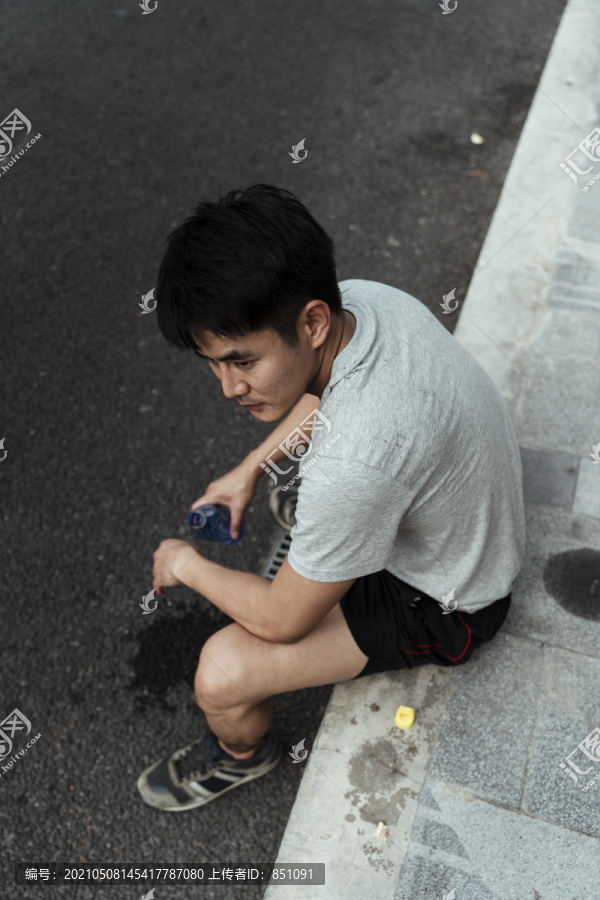 顶视图：亚洲年轻人坐在路边，手里拿着一瓶水。