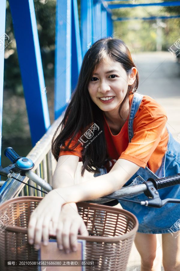一个长发女孩穿着橙色t恤和牛仔裤，在蓝色的桥上弯腰骑着自行车。