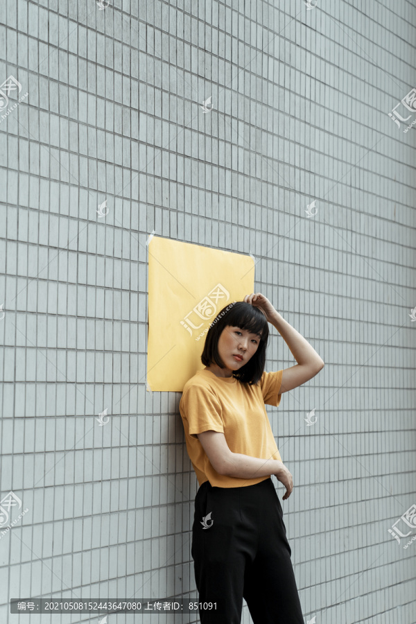 年轻的泰国亚裔女子深色短发穿着橘色衬衫站在街上，头后贴着墙纸。