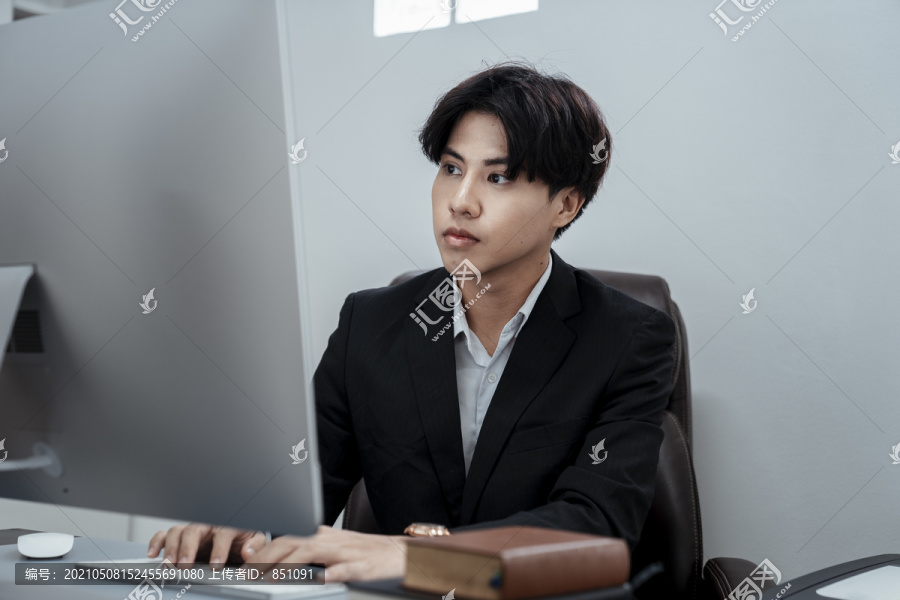 穿着黑色西装的亚洲商人坐在办公室的椅子上用电脑工作。