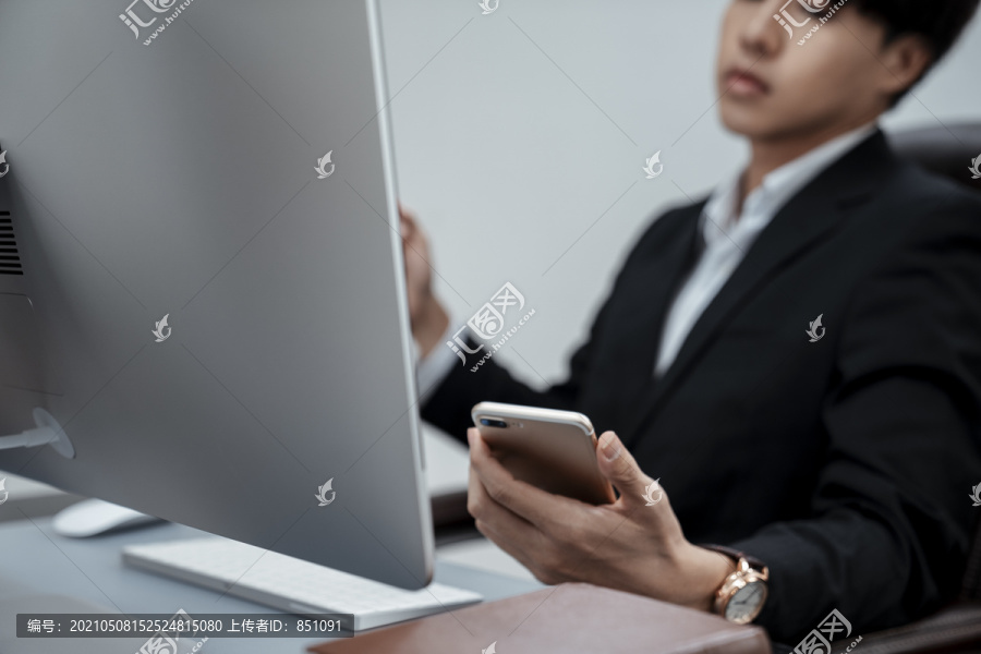 穿着黑色西装的亚洲商人在使用数字智能手机的同时使用笔记本电脑、查看移动应用程序或浏览互联网、在现代办