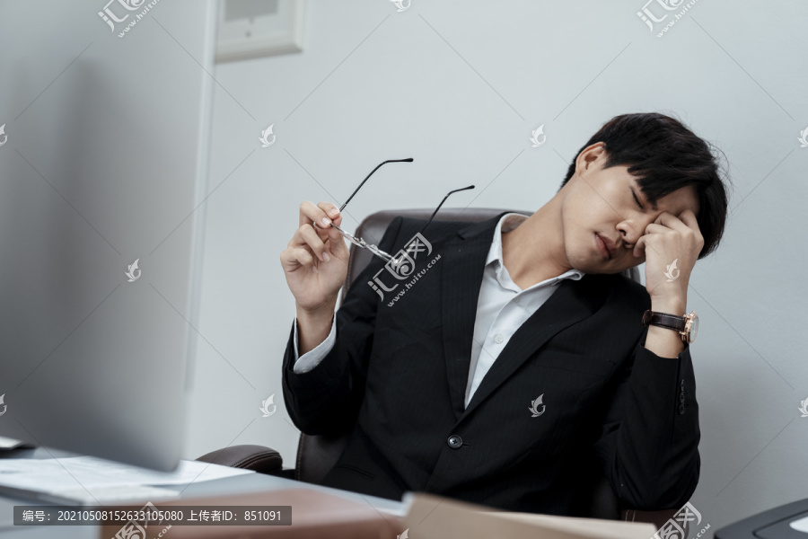疲惫的亚洲商人穿着黑色西装坐在办公桌的椅子上。