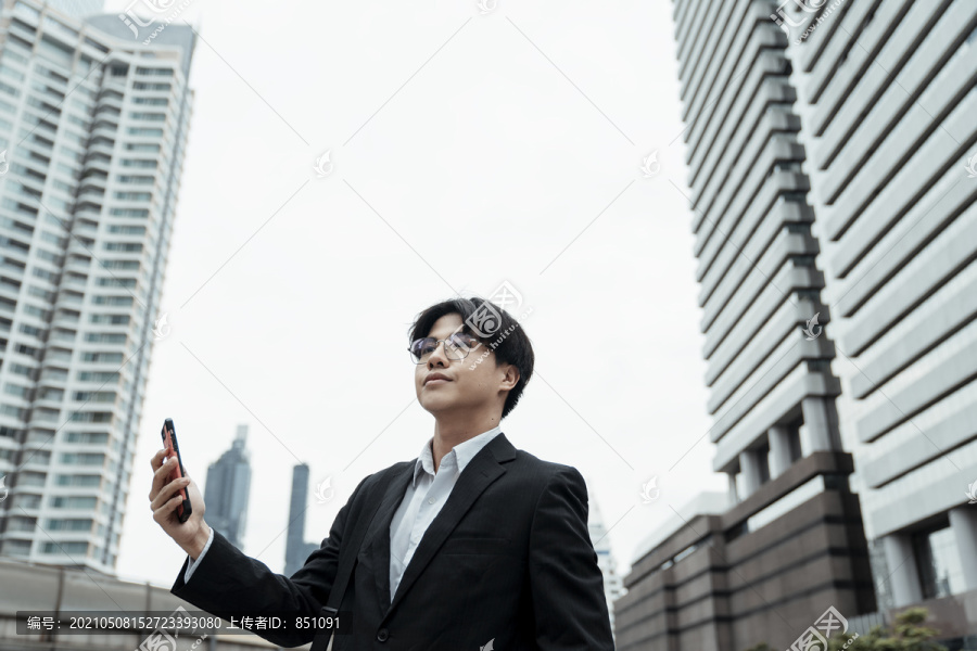 年轻聪明的亚洲商务人士身穿现代黑色西装，以手机智能手机为背景，在建筑城市中成功地实现了互联网网络连接