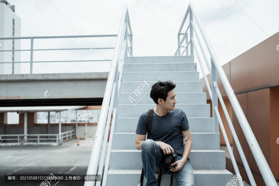 年轻英俊的亚洲摄影师坐在楼梯上拿着相机在屋顶拍照。