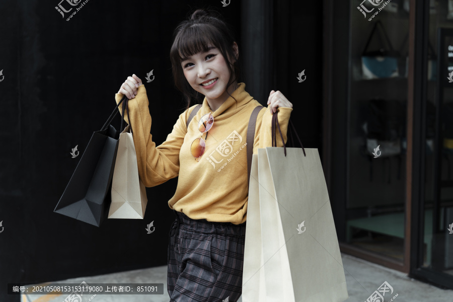兴高采烈的亚洲年轻女子穿着黄色毛衣，拿着许多纸袋在购物中心外散步。