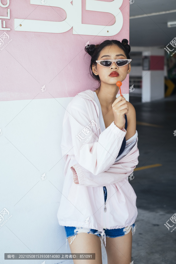 年轻迷人的亚泰女性深色双髻发型，粉色头巾，戴着时尚眼镜，手里拿着棒棒糖在停车场。