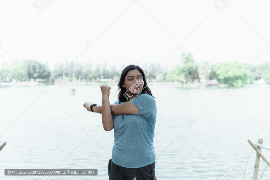 在公园的湖边，胖乎乎的亚洲女子穿着蓝色运动服伸展手臂。