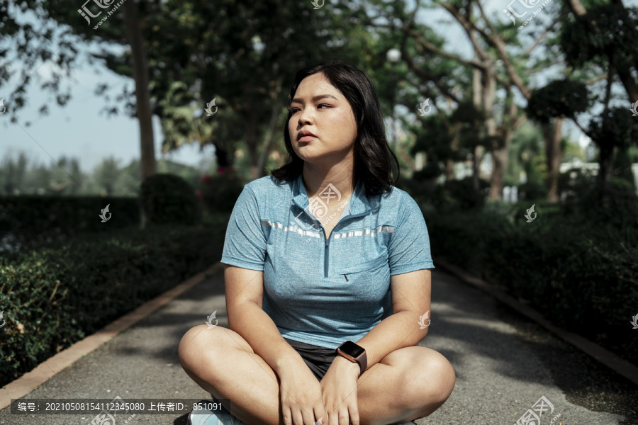 胖乎乎的运动型亚洲女人，穿着蓝色运动服，坐在跑道上休息。厌倦了跑步。