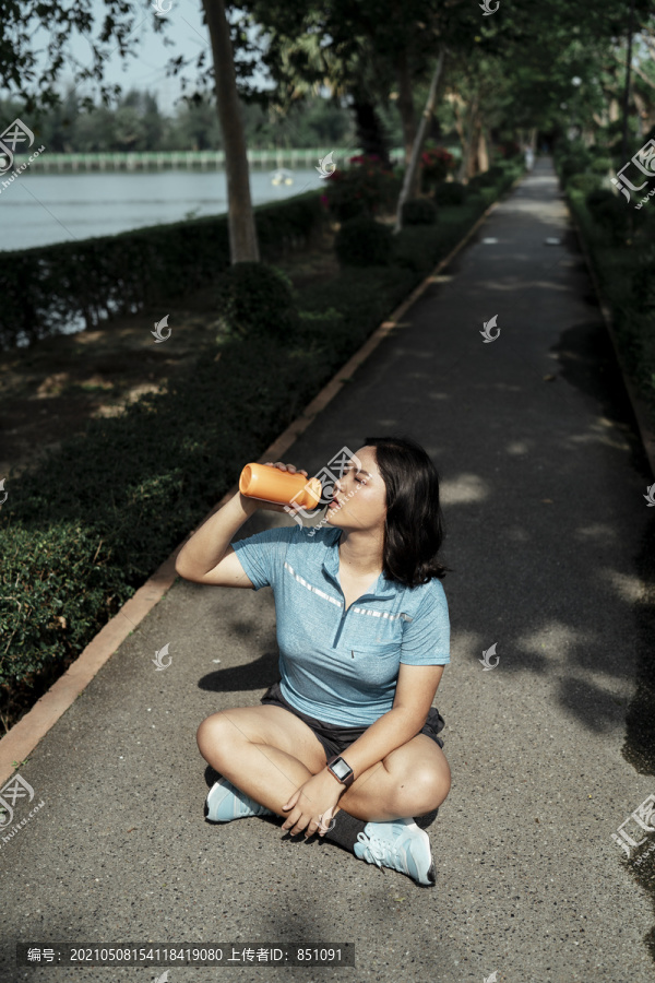 胖乎乎的运动型亚洲女子，穿着蓝色运动服，坐在跑道上，在公园喝着瓶装水。