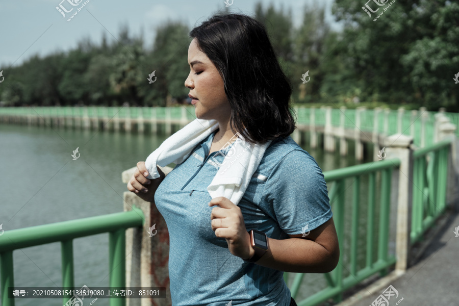 胖乎乎的运动型亚洲女性，穿着蓝色运动服，脖子上挂着白色毛巾，跑步后放松。