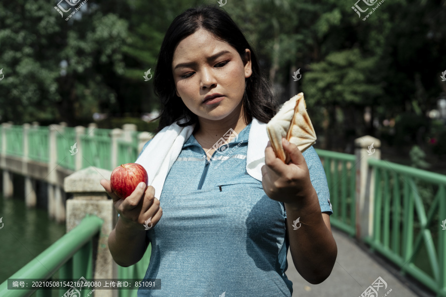 艰难的选择-胖乎乎的运动型亚洲女人穿着蓝色运动服选择苹果和三明治。健康的或不健康的。