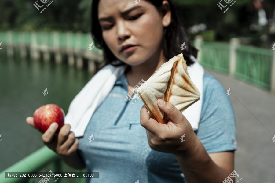 艰难的选择-胖乎乎的运动型亚洲女人穿着蓝色运动服选择苹果和三明治。健康的或不健康的。