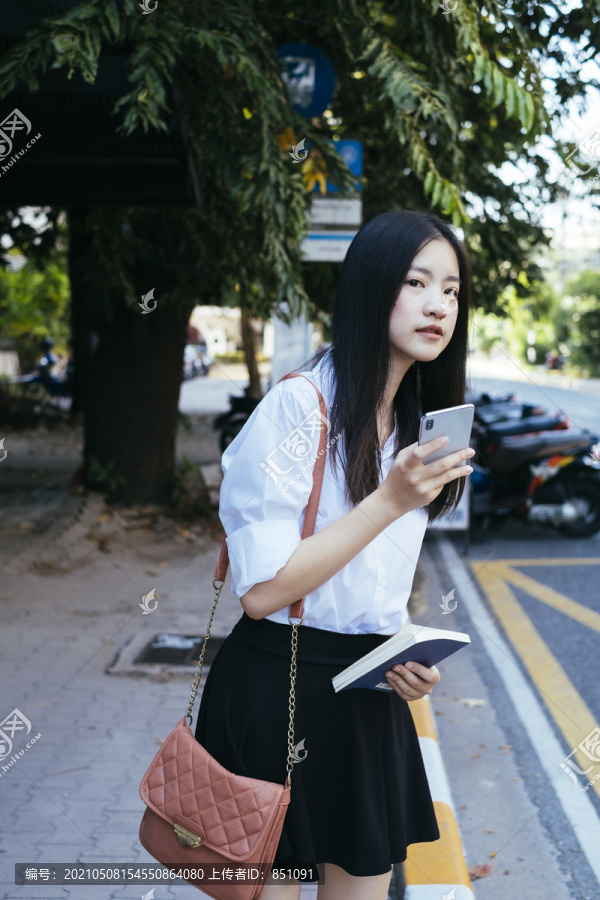 美丽的黑发青年大学生身着校服手持智能手机在人行道上等待交通工具。