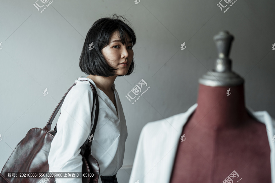 年轻的亚洲设计师短发女子拎着皮包，在展厅里看着自己戴在假人上的布料。